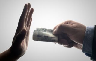 Slika prikazuje eno roko, ki daje zvitek denarja, in drugo roko, ki zavrača denar.