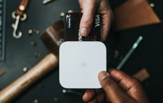 Slika prikazuje dve roki. Ena roka drži pos terminal za plačila, druga pa drži črno Visa kartico, s katero plačuje.