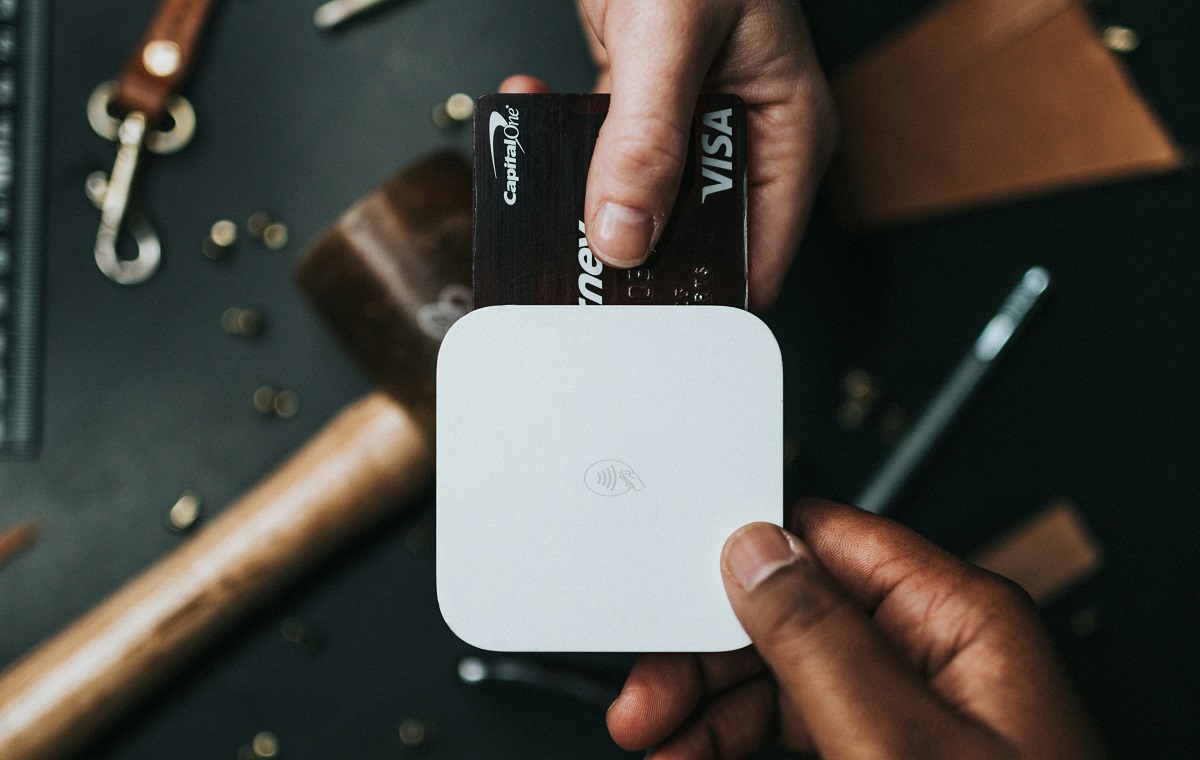 Slika prikazuje dve roki. Ena roka drži pos terminal za plačila, druga pa drži črno Visa kartico, s katero plačuje.