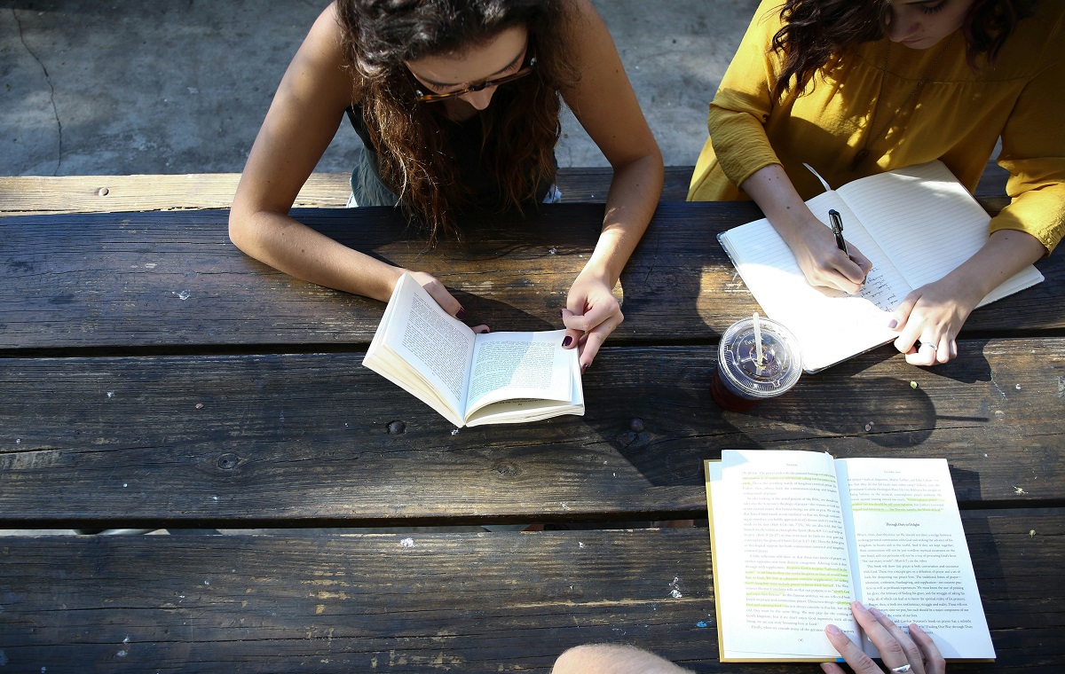 Slika prikazuje dve osebi ženskega spola z rjavimi lasmi. Sedita za mizo in imata v rokah zvezke.