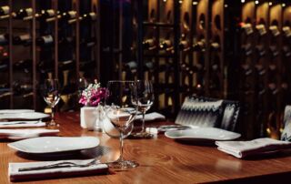 Slika prikazuje zatemnjeno restavracijo in mize, na katerih so pogrinjki, v ozadju pa steklenice na policah.