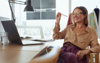 Slika prikazuje mlado poslovno žensko, ki je oblečena v rjavo srajco in vijolične hlače. Sedi za rjavo pisalno mizo, na kateri je prenosni računalnik in črna pisalniška luč. V ozadju so okna in obešalniki z oblačili.