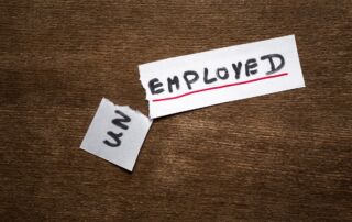 Slika prikazuje list papirja, na katerem piše Unemployed. List je raztrgan pri preponi Un. List je na rjavi leseni podlagi.