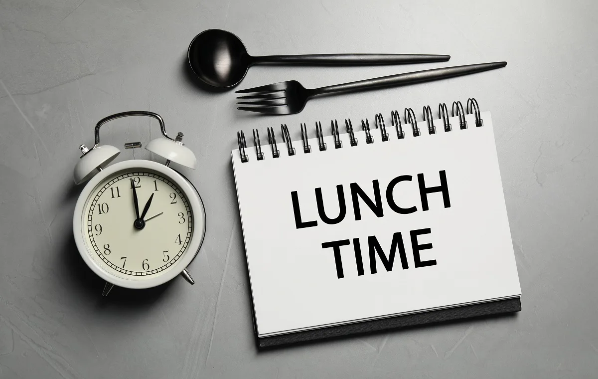 Zvezek z besedno zvezo Lunch Time, jedilni pribor in budilka na svetlo sivi mizi.