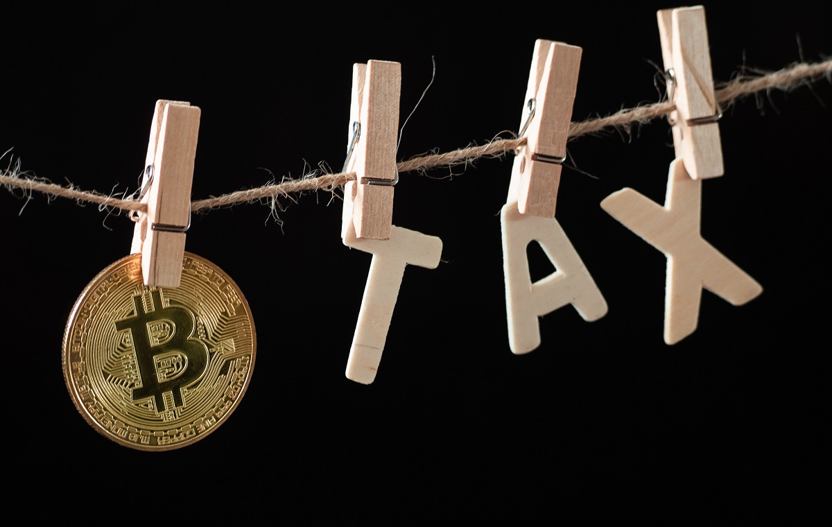 Zlati kovanec z logotipom Bitcoina in črkami, ki sestavljajo besedo 'TAX', obešenimi na vrvici s kljukicami. Slika simbolizira obdavčitev kriptovalut.